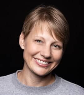 Headshot of a middle aged Caucasian woman smiling. She has short blonde hair and is wearing a grey shirt